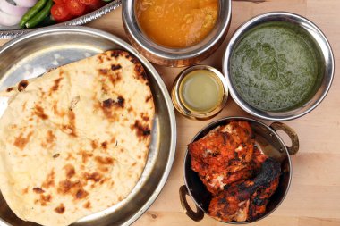 Baharatlı kırmızı tavuk ızgara tikka tandır nan ekmeği yeşil ıspanak körili yoğurt sosu domates salatalıklı soğan salatası metal paslanmaz çelik pirinç pirinç pirinç bakır kap hafif ahşap zemin üzerinde