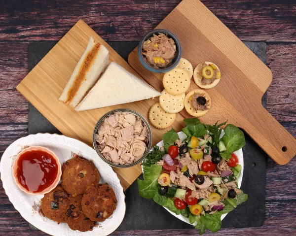 Tin can tuna fish meat chunk flake product recipes salad spread topping tapas cracker sandwich patty burger kebab on wooden black slate board over rustic wooden table