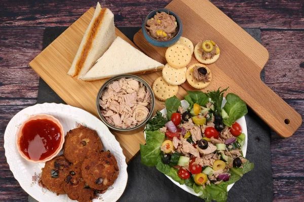 Tin can tuna fish meat chunk flake product recipes salad spread topping tapas cracker sandwich patty burger kebab on wooden black slate board over rustic wooden table