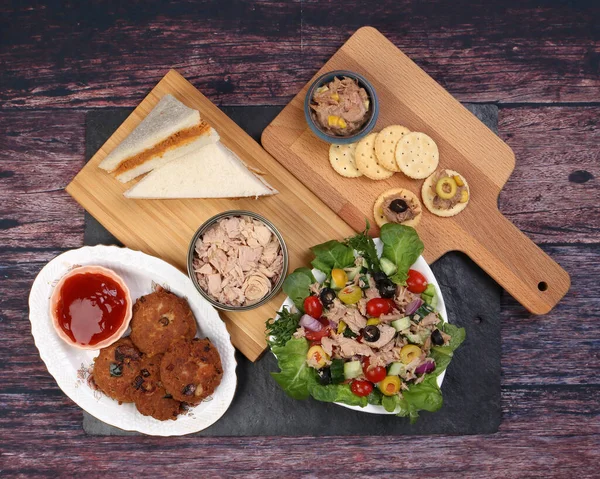 Tin can tuna fish meat chunk flake product recipes salad spread topping tapas cracker sandwich patty burger kebab on wooden black slate board over rustic wooden table