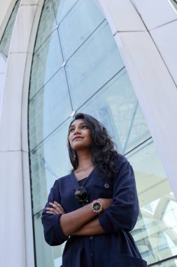 Koyu mavi eşofmanlı genç, çekici Hintli kadın uzun cam bir kentin önünde kendinden emin bir şekilde çapraz koluna bakar.