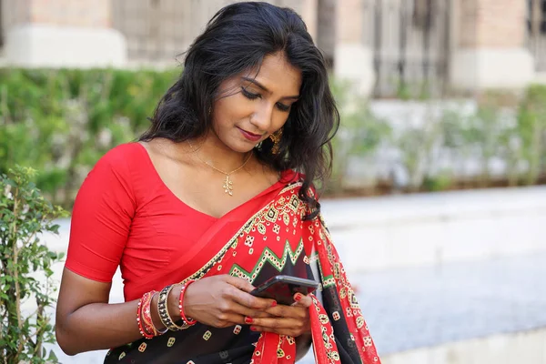 Young attractive beautiful Indian woman in red and black saree texting reading massage smartphone outdoors urban brick wall background