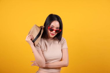 Güzel, genç, Güneydoğu Asyalı kadın kırmızı çerçeveli güneş gözlüklü şapka moda zirvesi sarı turuncu arka planda elinde şapkayla mutlu bir gülümseme.