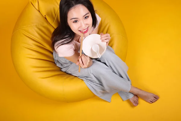 Beautiful young south east Asian woman holding empty chopstick soup spoon bowl posing see eat taste smell feed offer satisfaction yummy siting on beanbag seat yellow orange background top view