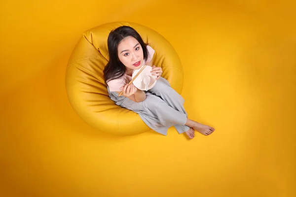 Beautiful young south east Asian woman holding empty chopstick soup spoon bowl posing see eat taste smell feed offer satisfaction yummy siting on beanbag seat yellow orange background top view