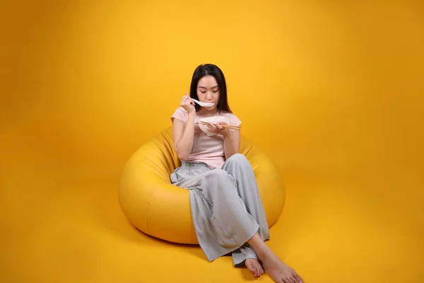 Beautiful young south east Asian woman holding empty chopstick soup spoon bowl pretend acting posing see eat taste smell feed offer satisfaction yummy siting on beanbag seat yellow orange background