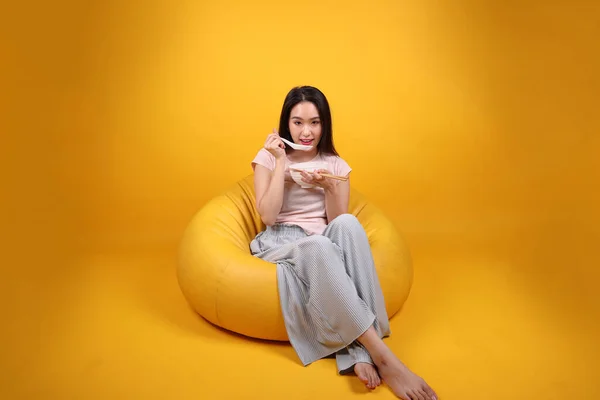 Beautiful young south east Asian woman holding empty chopstick soup spoon bowl pretend acting posing see eat taste smell feed offer satisfaction yummy siting on beanbag seat yellow orange background