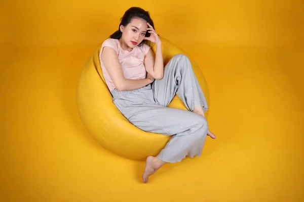 Beautiful young south east Asian woman sits on a yellow beanbag seat orange yellow color background pose fashion style elegant beauty mood expression rest relax think emotion sleep top view