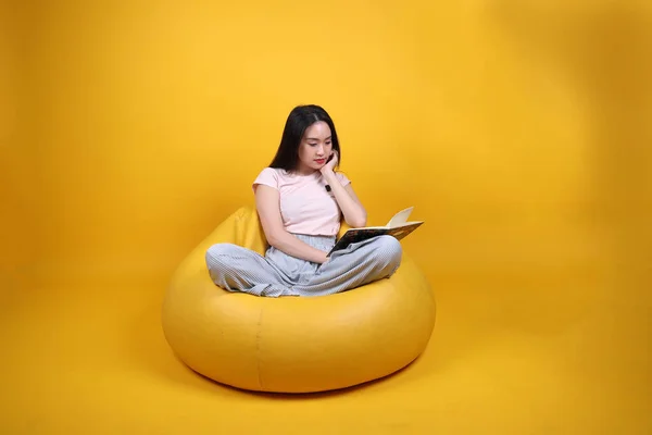 Beautiful young south east Asian woman sits on a yellow beanbag seat orange yellow color background pose fashion style elegant beauty mood expression rest relax read book think