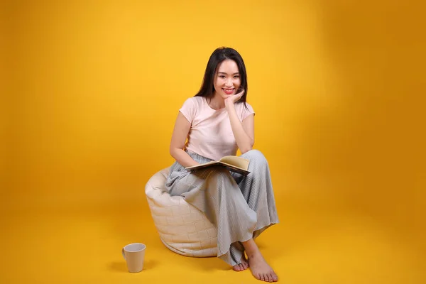 Beautiful young south east Asian woman sits on a white beanbag seat orange yellow color background pose fashion style elegant beauty mood expression relax smile look read book