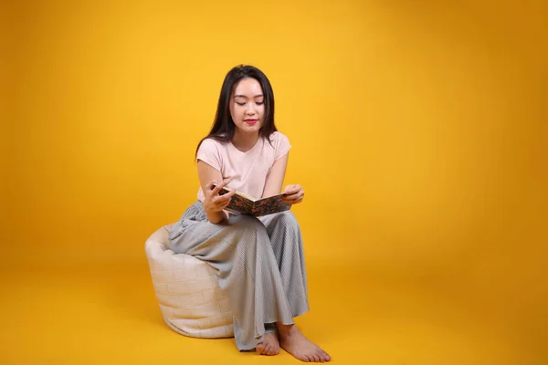 Beautiful young south east Asian woman sits on a white beanbag seat orange yellow color background pose fashion style elegant beauty mood expression relax smile look read book