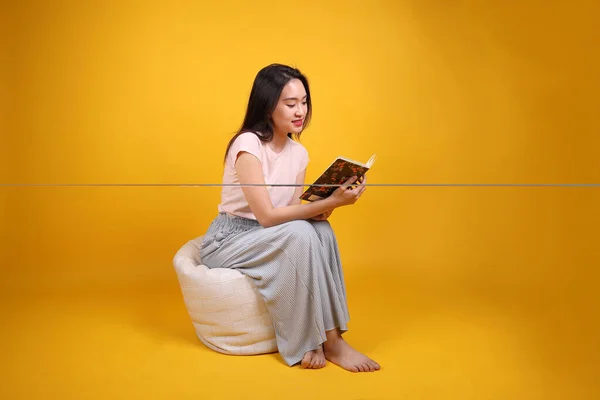 Beautiful young south east Asian woman sits on a white beanbag seat orange yellow color background pose fashion style elegant beauty mood expression relax smile look read book