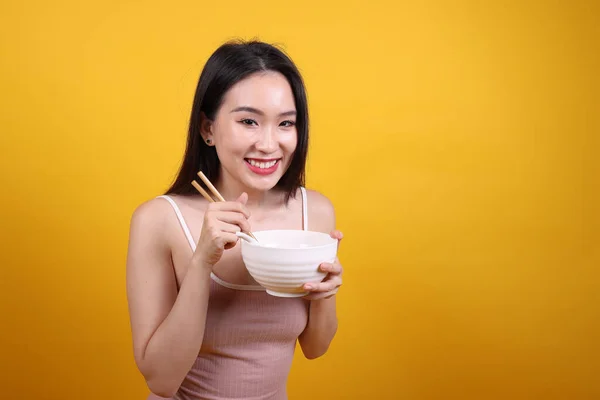 Beautiful young south east Asian woman holding chines empty chopstick Chinese soup spoon bowl utensil pretend acting posing see eat taste smell feed offer satisfaction yummy yellow orange background