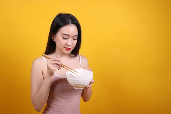 Beautiful young south east Asian woman holding chines empty chopstick Chinese soup spoon bowl utensil pretend acting posing see eat taste smell feed offer satisfaction yummy yellow orange background