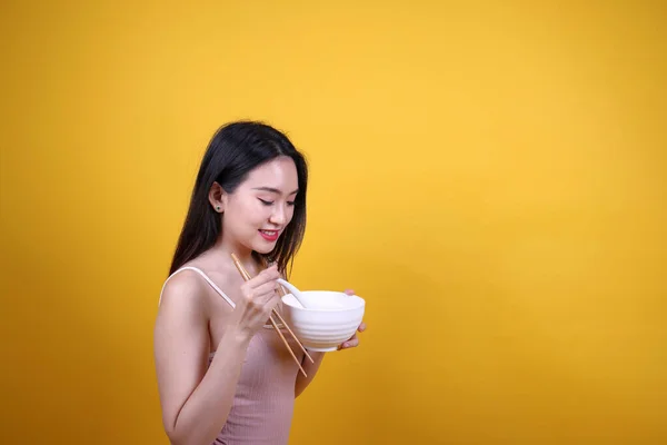 Beautiful young south east Asian woman holding chines empty chopstick Chinese soup spoon bowl utensil pretend acting posing see eat taste smell feed offer satisfaction yummy yellow orange background