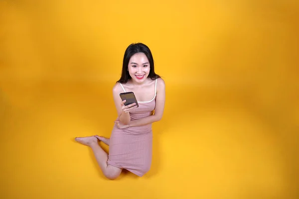 Beautiful young south east Asian woman sit down on floor yellow orange color background hold use text smart phone pose fashion style elegant beauty mood look at camera