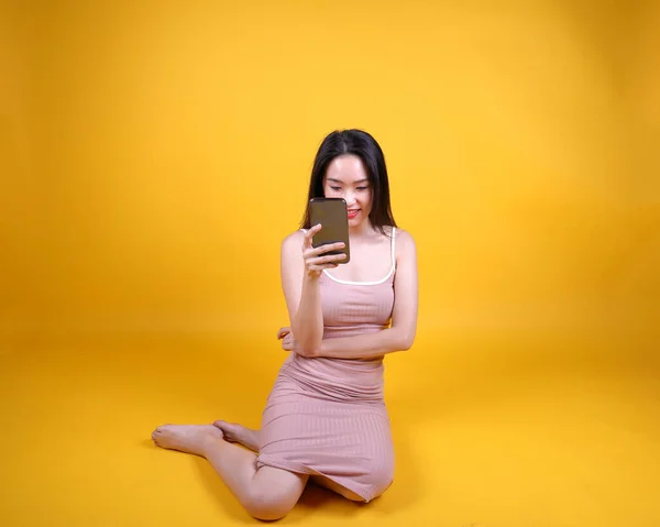 Beautiful young south east Asian woman sit down on floor yellow orange color background hold use text smart phone pose fashion style elegant beauty mood