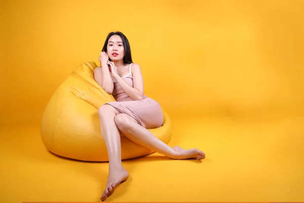 Beautiful young south east Asian woman sit on a yellow orange beanbag seat color background pose fashion style elegant beauty mood