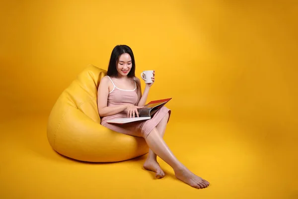 Beautiful young south east Asian woman sit on a yellow orange beanbag seat color background relax read study think book magazine hold white coffee cup