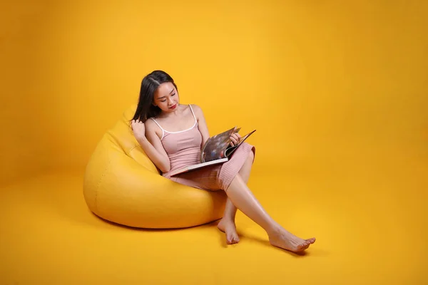 Beautiful young south east Asian woman sit on a yellow orange beanbag seat color background relax read study book magazine