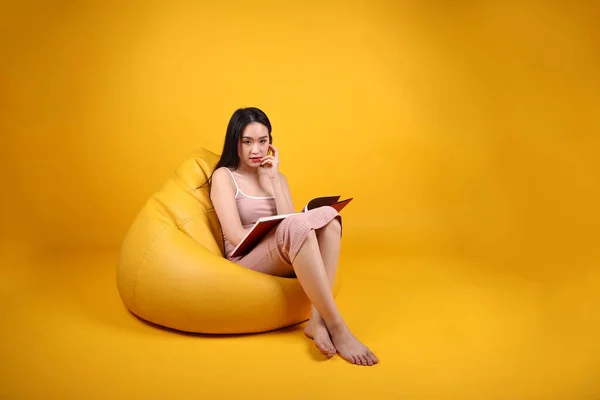 Beautiful young south east Asian woman sit on a yellow orange beanbag seat color background relax read study book magazine