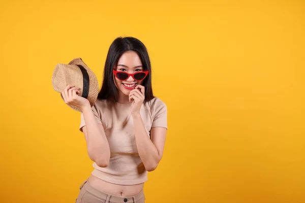 Beautiful young south east Asian woman red frame sunglass hat pose style fashion peak happy on yellow orange background holding hat in hand smile happy