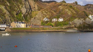 Barmouth, Galler, Mart 06, 2024: Sahil şeridi boyunca ilerleyen yolcu treni ön planda durgun su ve kayalık yamaçlara kurulmuş evler. Manzaralı bir Galler kıyı manzarası Demiryolu taşımacılığı ve denizcilik çevresinin birleşimi