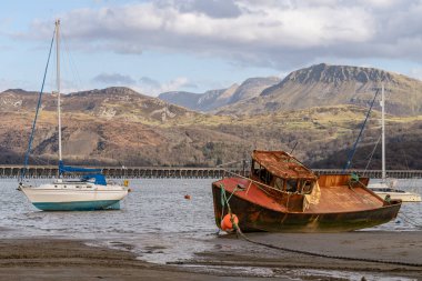 Barmouth, Galler, Mart 06, 2024: Paslı yelkenli ve modern yelkenli yatı limanda dinleniyor. Sakin kıyı suları, kumlu ön planlar ve arka plandaki dramatik Galler dağları çürüme ile boş zaman arasında çarpıcı bir tezat oluşturur.
