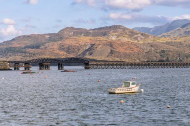 Barmouth, Galler, Mart 06, 2024: Arka planda tarihi demiryolu köprüsü ve dağlarla Mawddach Estuary 'de demirlemiş küçük bir balıkçı teknesi. Sakin bir kıyı manzarası deniz yaşamını ve Galler kırsalını gösteriyor.