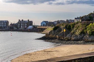 Barmouth, Galler, Mart 06, 2024, kıyı şeridi boyunca sakin kumlu sahil, deniz manzaralı taş binalar ve kayalıklar. Geleneksel bir Galler sahil kasabasının karakterini yansıtan sakin bir sahil sahnesi