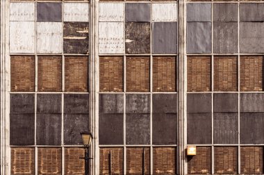 Birbirine uymayan ahşap ve metal panellerle tekrar tekrar kapatılmış sanayi pencereleri çürüme, terk edilme ve dayanıklılık gösteriyor. Ön cephe dokuyu, simetriyi ve yapı ile bozulma arasındaki zıtlığı vurgular.