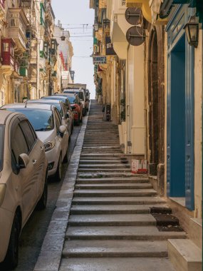 Valletta, Malta - 8 Nisan 2024: Valletta 'da park halindeki araçlar ve kentin dik topoğrafyası ve yoğun kentsel yapısını gösteren geleneksel kireçtaşı binalarla dolu dar bir sokak.