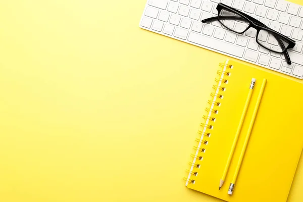 Student or freelancer workspace with keyboard, glasses, notepad, pencil on yellow background, flat lay. Space for text