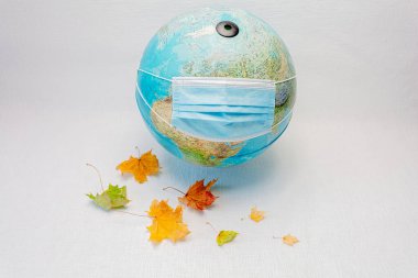 Globe tıbbi maske takıyor ve beyaz arka planda akçaağaç yaprakları var. Kavram, dünyamıza koronavirüs bulaştı. Autumn, karantinada evde kal.