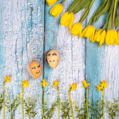 Sarı çiçekler ve yumurtaların bahar kompozisyonu mavi arka planda boyalı şirin bir yüz. Paskalya için tebrikler. Acil