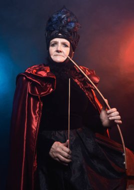 Muhteşem bir yaratık, tarihi bir karakter, bir fantezi kraliçesi, bir cadı, elinde bir yayla muhteşem bir kostüm içinde. Kadın, 60 'ından sonra model, stüdyoda, koyu arka planda.