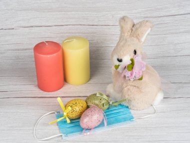 Tavşanın etrafında renkli yumurtalar ve mumlar. Mutlu Paskalyalar. Tavşan ve tıbbi maske, koronavirüs saati 19. Sağlık, tatil, maske güvende kalsın. Bahar sezonu. Mutlu Paskalya bayrağı ışık arkaplanı.