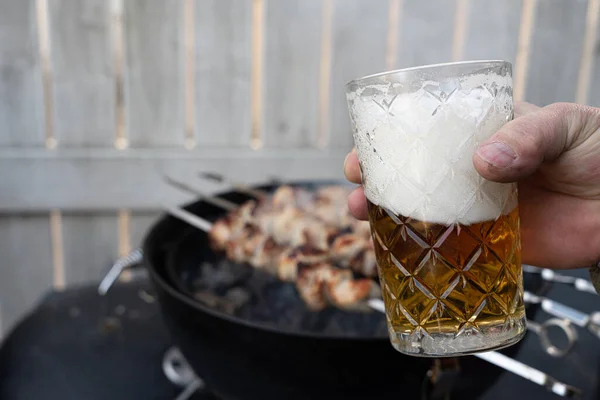 Glass of beer in hand on the background of a barbecue. Cooking blue pieces of meat on the grill. Traditional weekend picnic on a festive sunny day, harsh shadows. Street food.