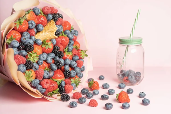 Edible bouquet of fresh berries, blueberries, raspberries, strawberries, hedgehog on a pink background with scattered berries and a jar for smoothies. View from the top .Close-up .Concept of a useful gift, congratulations on the holiday.