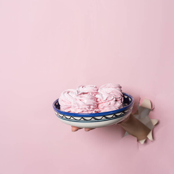 Hands hold a plate with homemade pink marshmallows through a torn hole on a pink paper background. Bakery advertising concept, recipe.