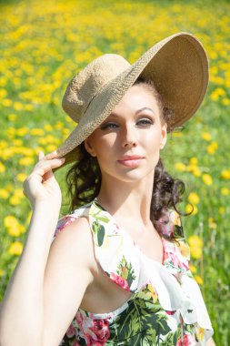 Hasır şapkalı bir kadın, esmer, karahindiba arka planında. Kameraya bakıyor. Yaz, güneşli gün.