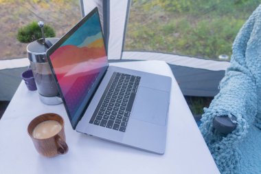 Masadaki çadır bir dizüstü bilgisayar, bir kahve demliği, bir fincan kahve. Günaydın konsepti. Doğada yaz tatili.