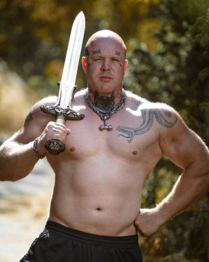 Espoo, Finlandiya, 25 Temmuz 2021. Dövmeli, atletik, vücut geliştiren, çıplak gövdeli, arka planda dövüş kılıcı olan bir adam..