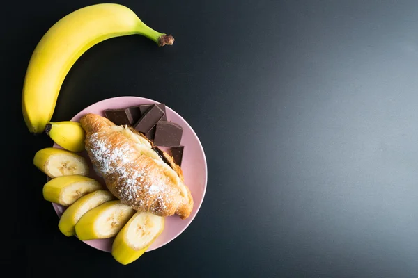 Свіжий круасан з шоколадом, банан на рожевій тарілці на чорному ба — стокове фото