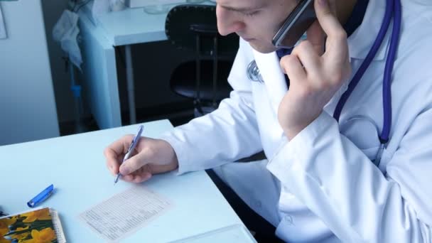 Jovem médico caucasiano falando por telefone e escrevendo prescrição médica no hospital — Vídeo de Stock