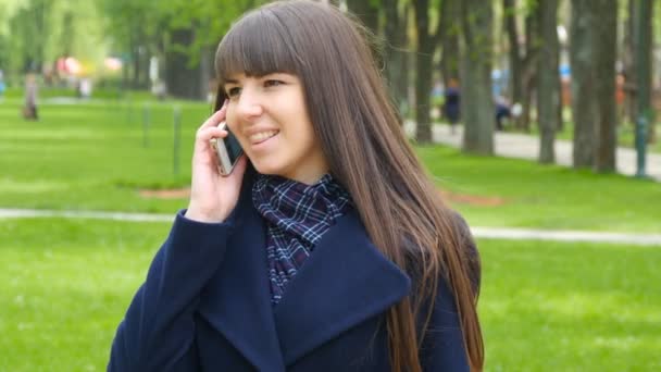 Una joven hablando por teléfono móvil en un parque de la ciudad. Verde hierba de primavera agradable de jardín en el fondo. Mujer de negocios hablando y sonriendo. Primer plano — Vídeos de Stock