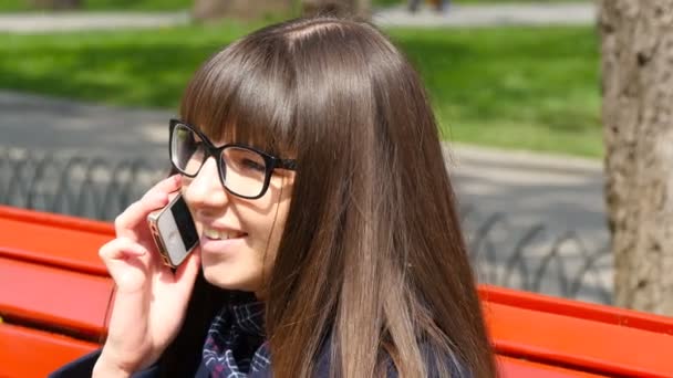 Genç kadın bir şehir parkı modile telefonda konuşurken gözlük. Bahar açık kırmızı bir Bank üzerinde oturan ve Smartphone'da konuşan kız. Yakın çekim — Stok video