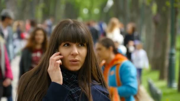 Atractiva joven con abrigo azul hablando por teléfono móvil en el callejón del parque. Chica feliz hablando en smartphone, al aire libre. Primer plano — Vídeo de stock