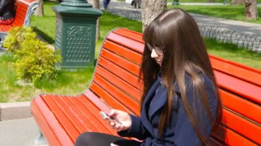 Gözlüklü genç kadın hücre smartphone bir şehir parkında kullanır. Bir kırmızı üzerinde oturan kız baharda açık tezgah ve bir cep telefonu kullanır. Yakın çekim