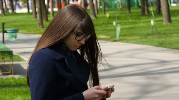 Bella donna in occhiali utilizza smartphone cellulare all'aperto nel parco dettaglio. Giovane ragazza felice attraente si rilassa in un parco cittadino e utilizza un telefono cellulare. Sembra molto felice e contenta. — Video Stock